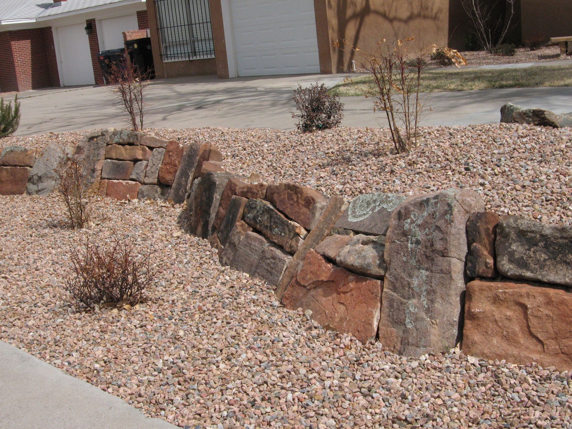 A low retaining wall made of reddish-brown rocks in a gravel bed with sparse shrubs.