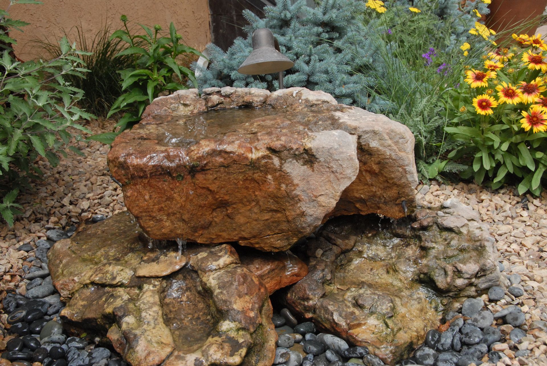 A cascading rock fountain in a garden setting with various flowers and plants.