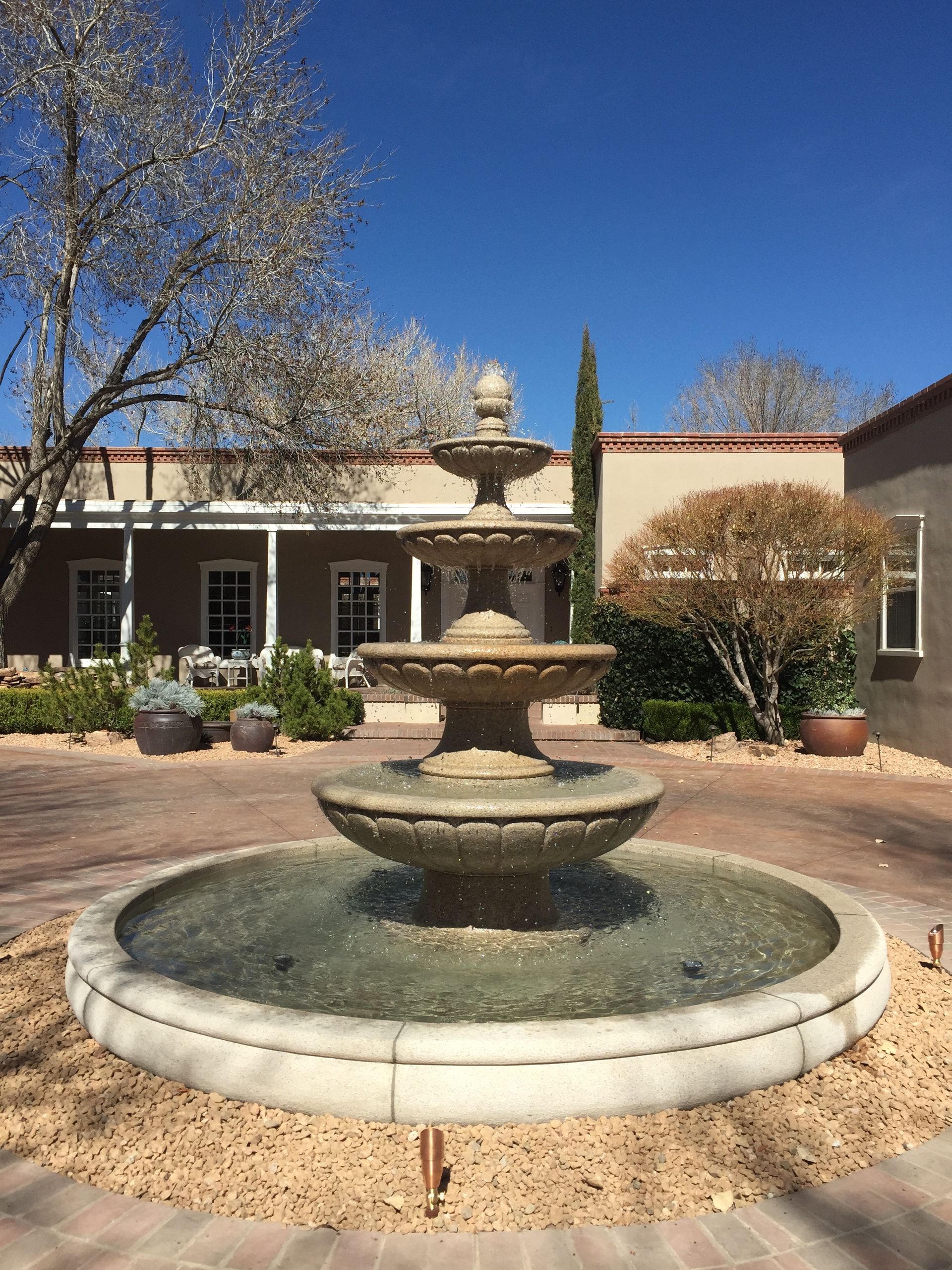 Stone fountain in a courtyard with blooming trees and a light brown building under a clear, blue sky.