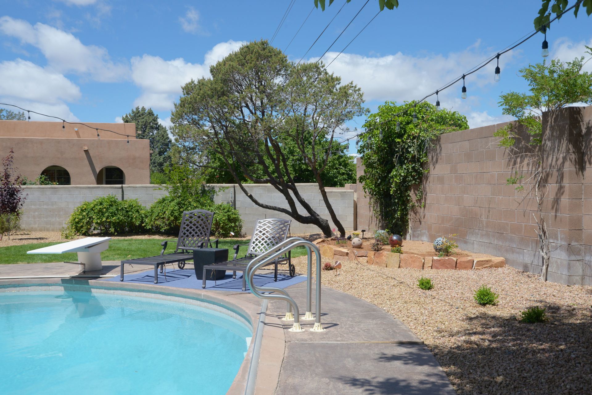 Swimming pool in a backyard with lounge chairs, a tree, and a wall.