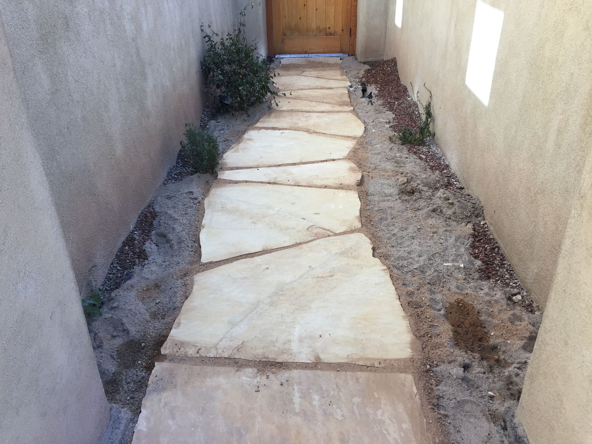 Stone path leads to a wooden door, bordered by beige walls and gravel with small plants.