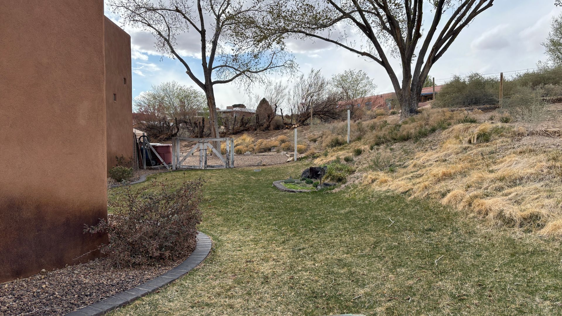 Grassy yard with brown stucco wall, gate, and dry brush, under a cloudy sky.