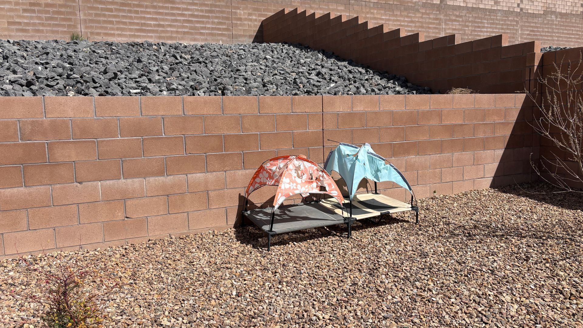 A baby stroller with two canopies, orange and blue, sits on gravel next to a brick wall.