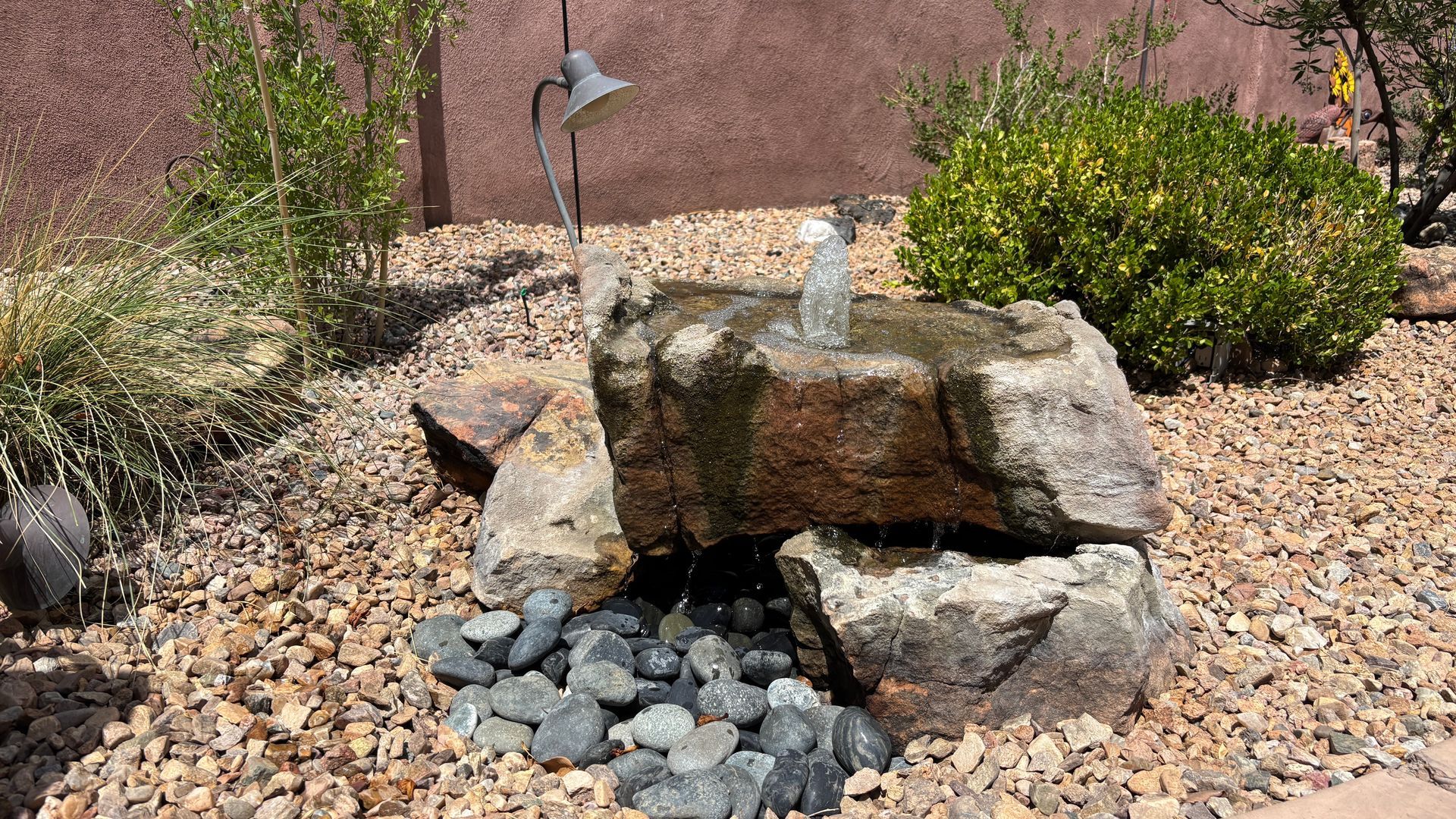 Stone fountain in a rock garden, water splashing. Brown gravel, rocks, and green bushes.