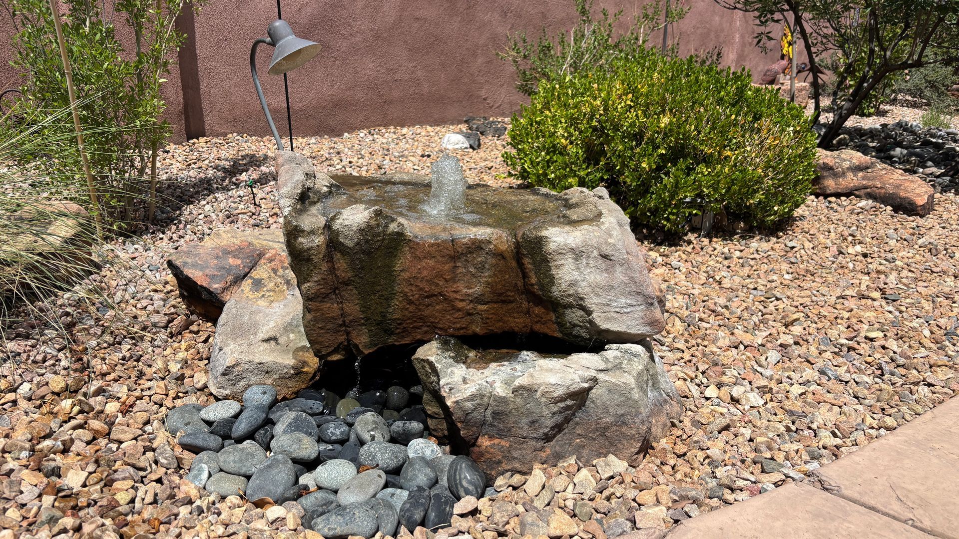Water fountain made of rocks with water flowing, surrounded by gravel and plants.