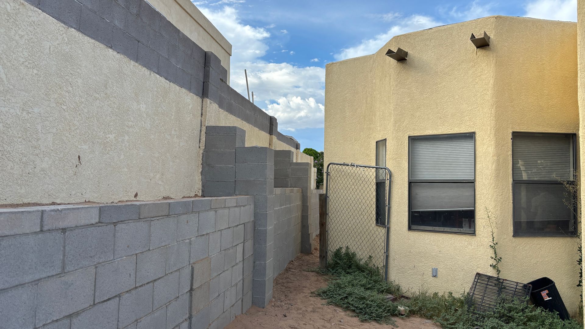 Narrow alley between stucco buildings with cinder block walls, blue sky visible.
