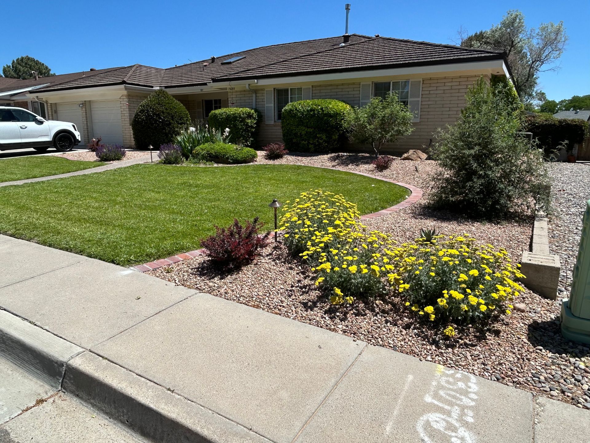 Low-angle view of a beige brick house with manicured lawn and flowerbeds, bordered by a concrete sidewalk.