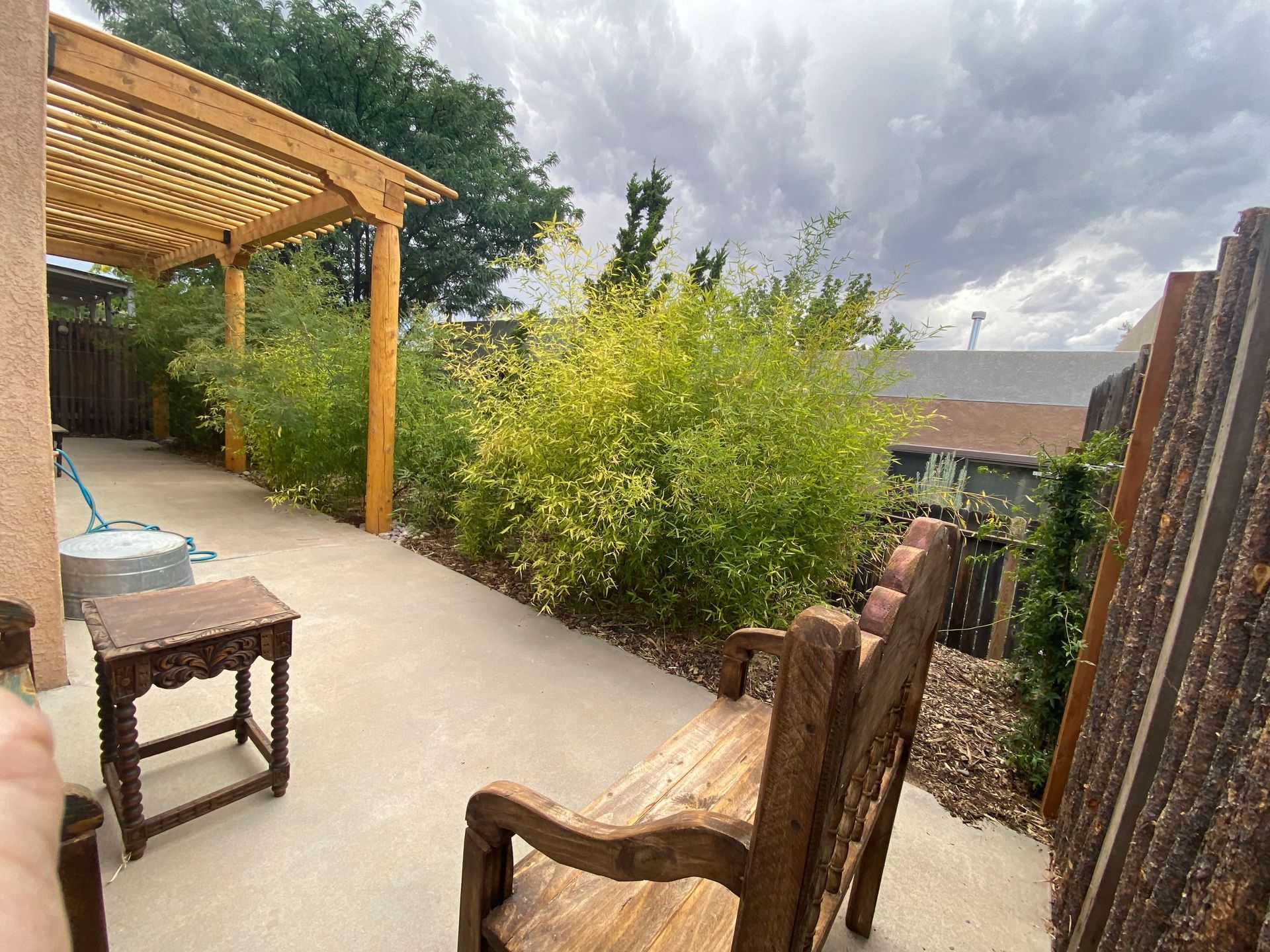 Wooden bench and small table on patio, with bamboo and pergola under cloudy sky.