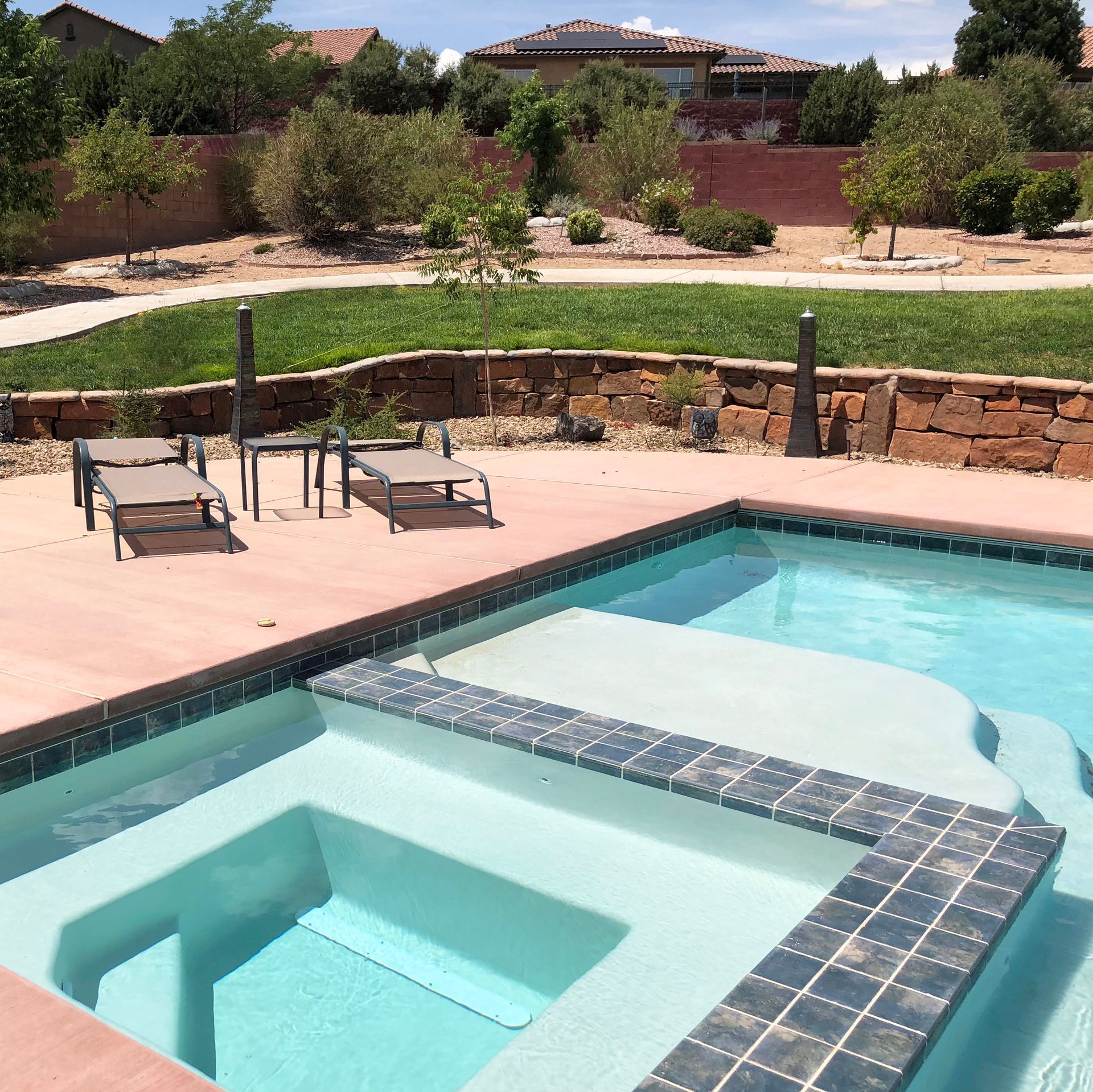 Swimming pool and hot tub with lounge chairs in a backyard on a sunny day.