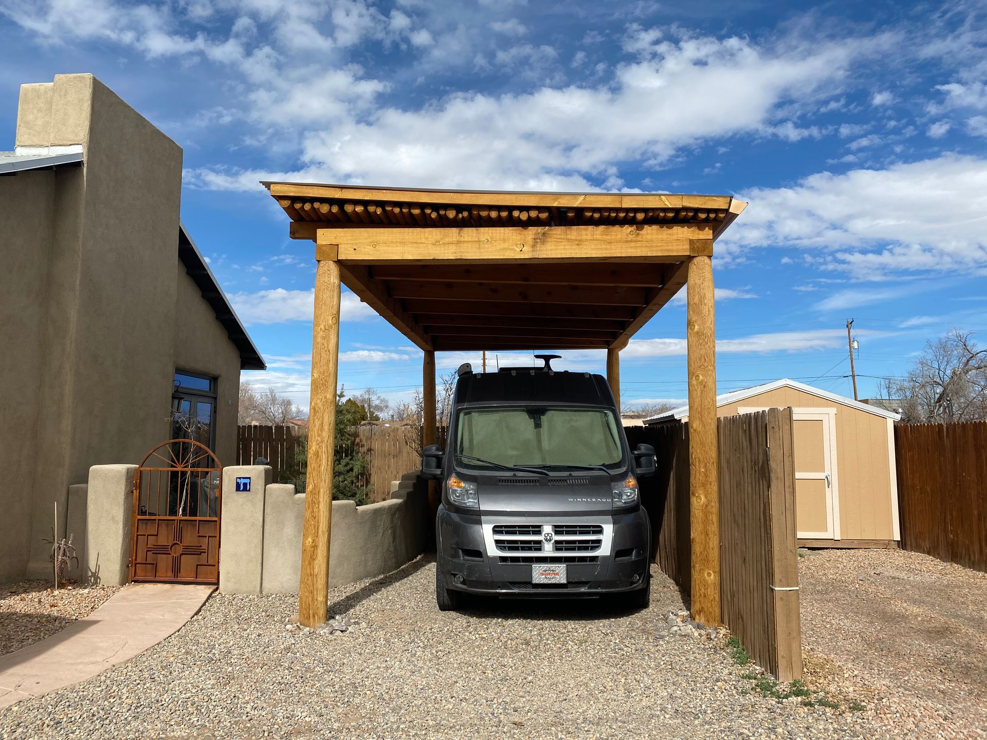 A gray van parked under a wooden carport in a gravel driveway.