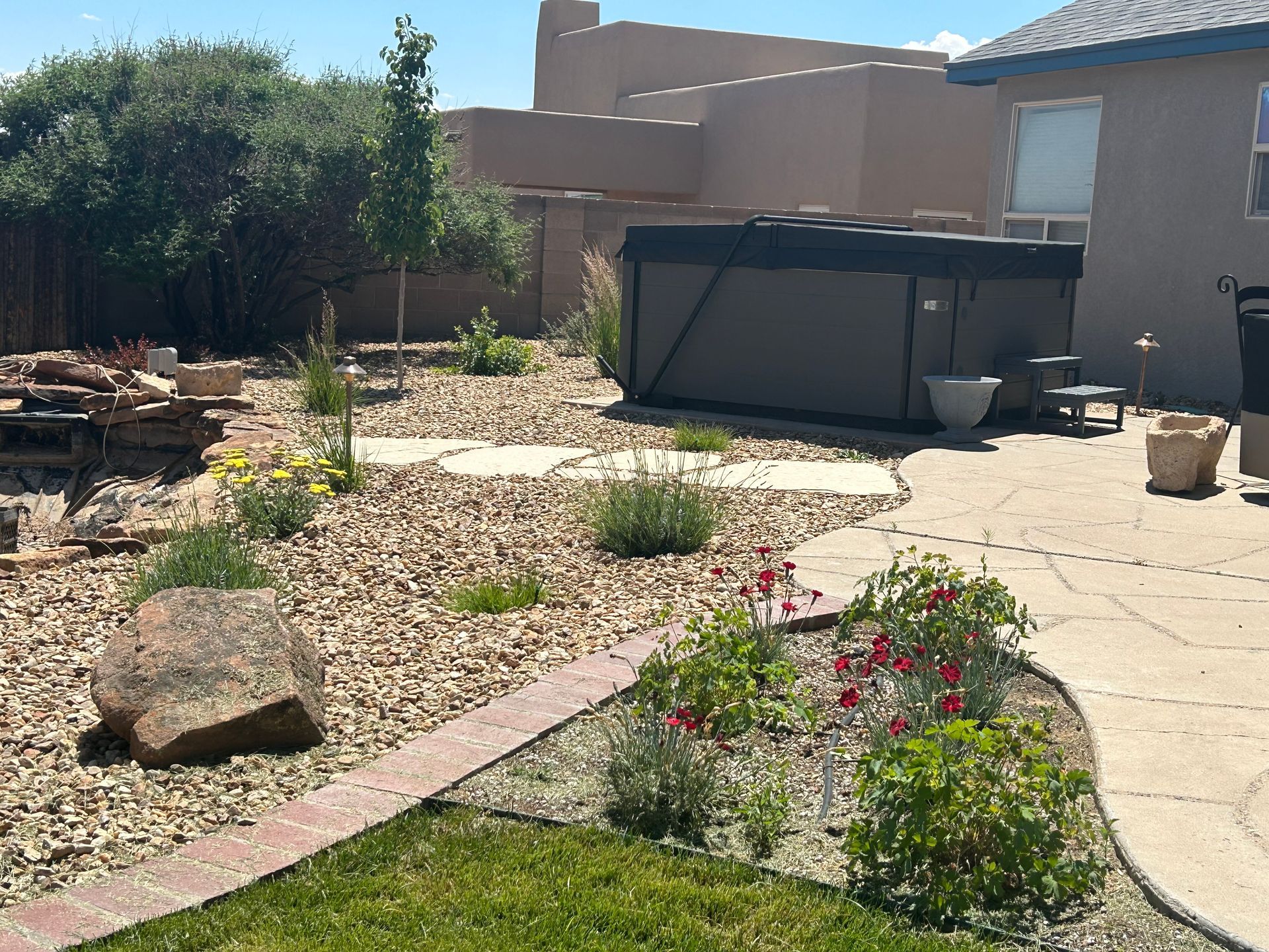 Backyard with a hot tub, stone pathway, rock bed, and plants. Beige stucco house in background.