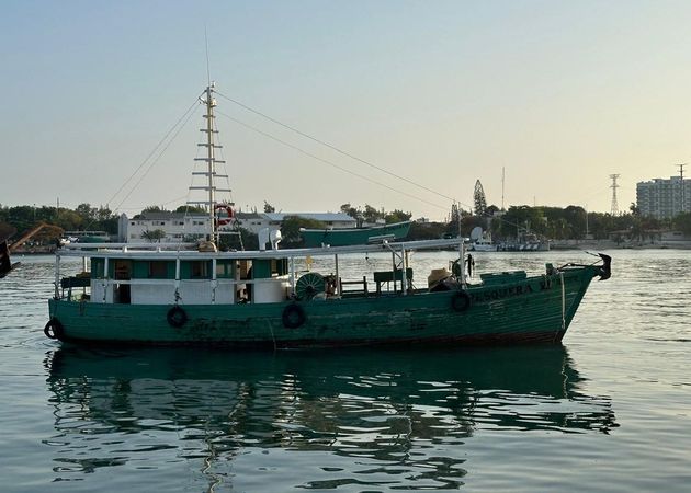 Green boat on calm water at sunset, with a tall mast and reflections on the harbor.