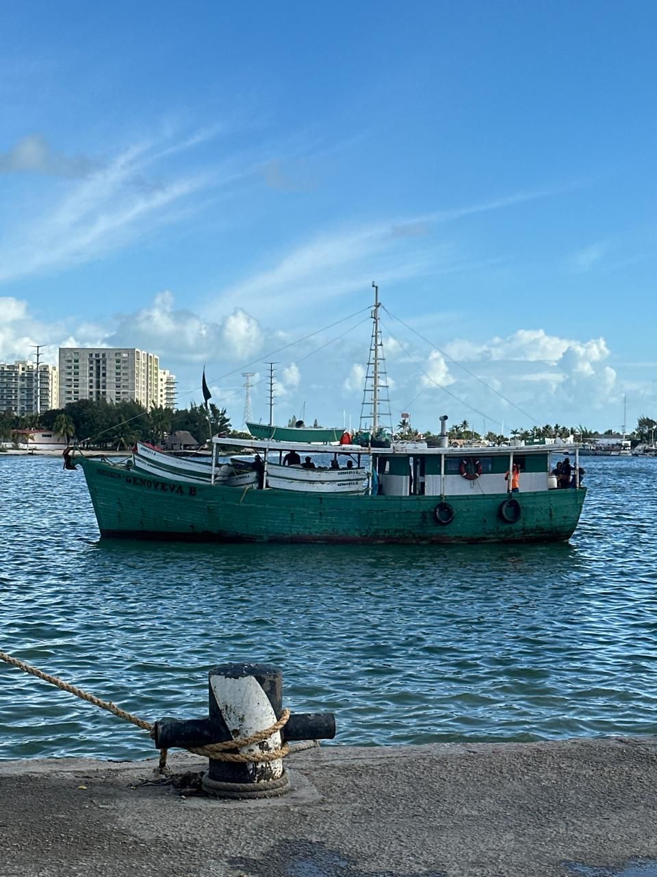 Green fishing boat on blue water near a city shoreline under a bright sky