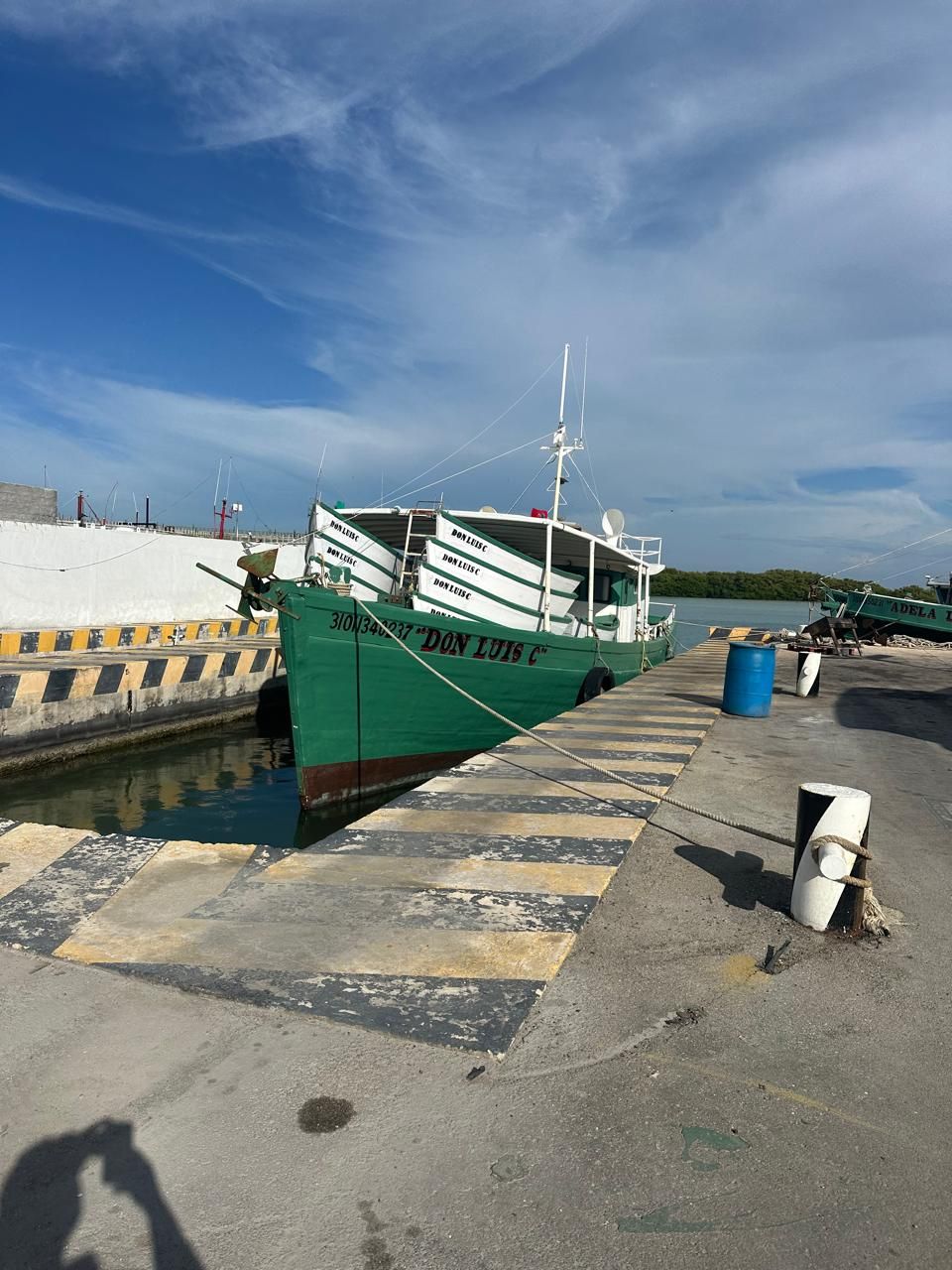 Green fishing boat docked at a harbor under a blue sky, with concrete pier and mooring posts nearby.