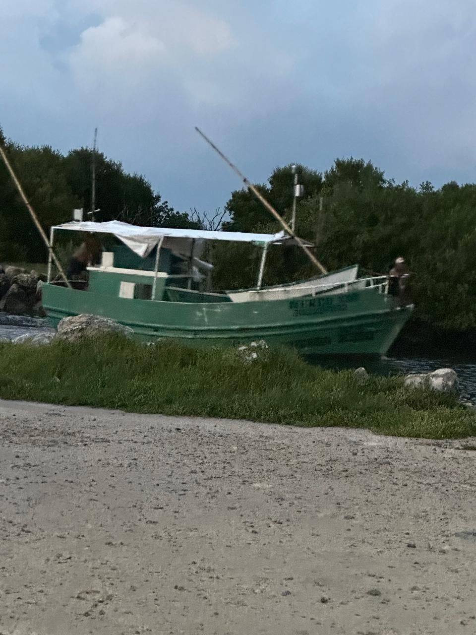 Green fishing boat on shore with raised masts and white canopy, near rocky water and trees under cloudy sky