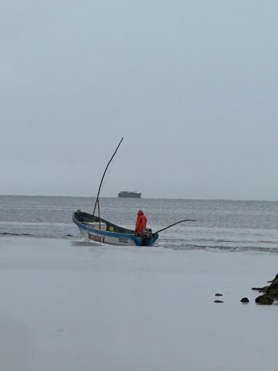 Small fishing boat with a person in a red jacket on a gray beach at low tide, with the sea in the background