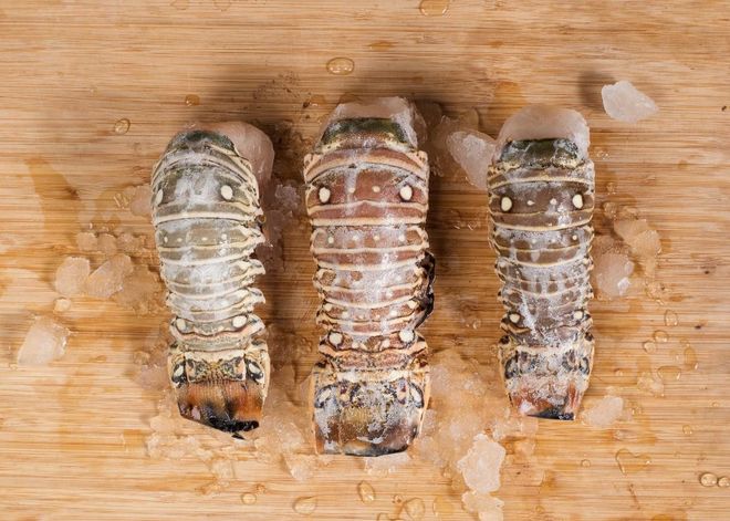 Three lobster tails on a wooden cutting board, lightly seasoned and dusted with salt.