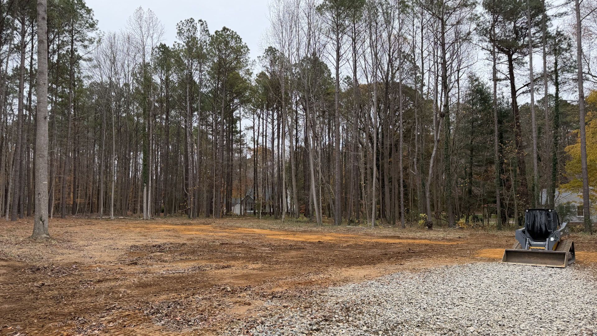 Clearing of land with gravel in the foreground, trees in the background, and a skid steer.