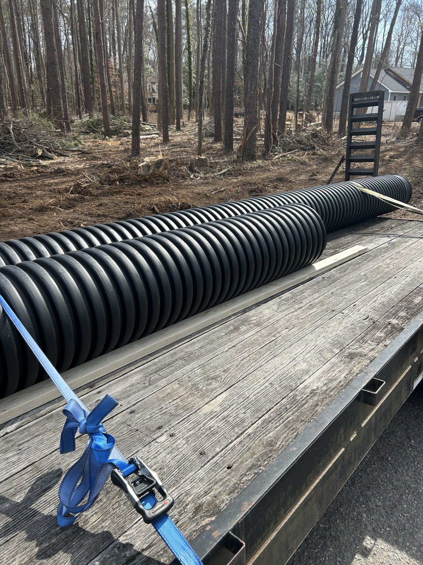 Black corrugated pipes loaded on a flatbed trailer in a wooded area. Blue straps secure the pipes.