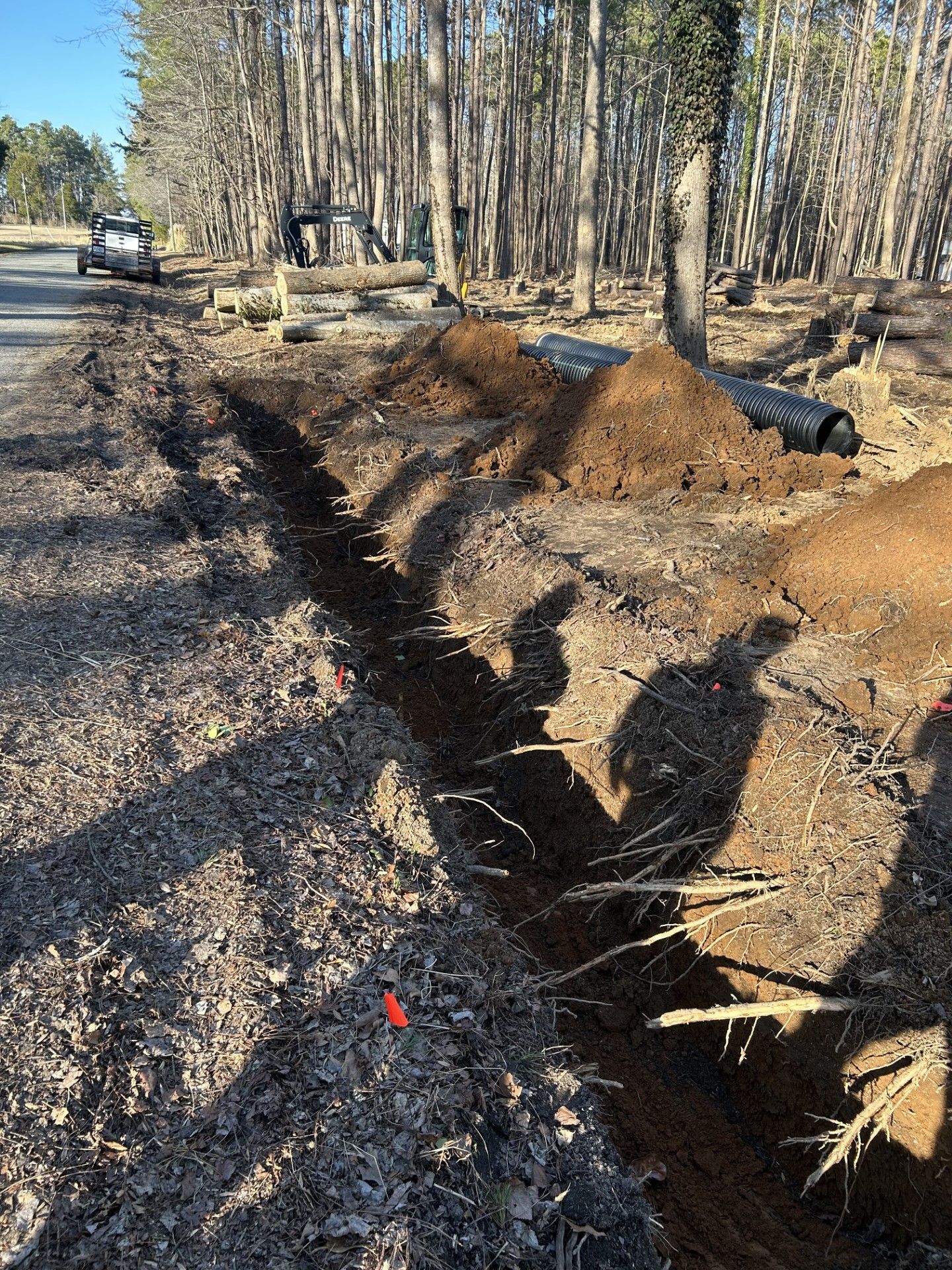Trench dug alongside a road, dirt piles, and a black pipe visible. Trees in the background.