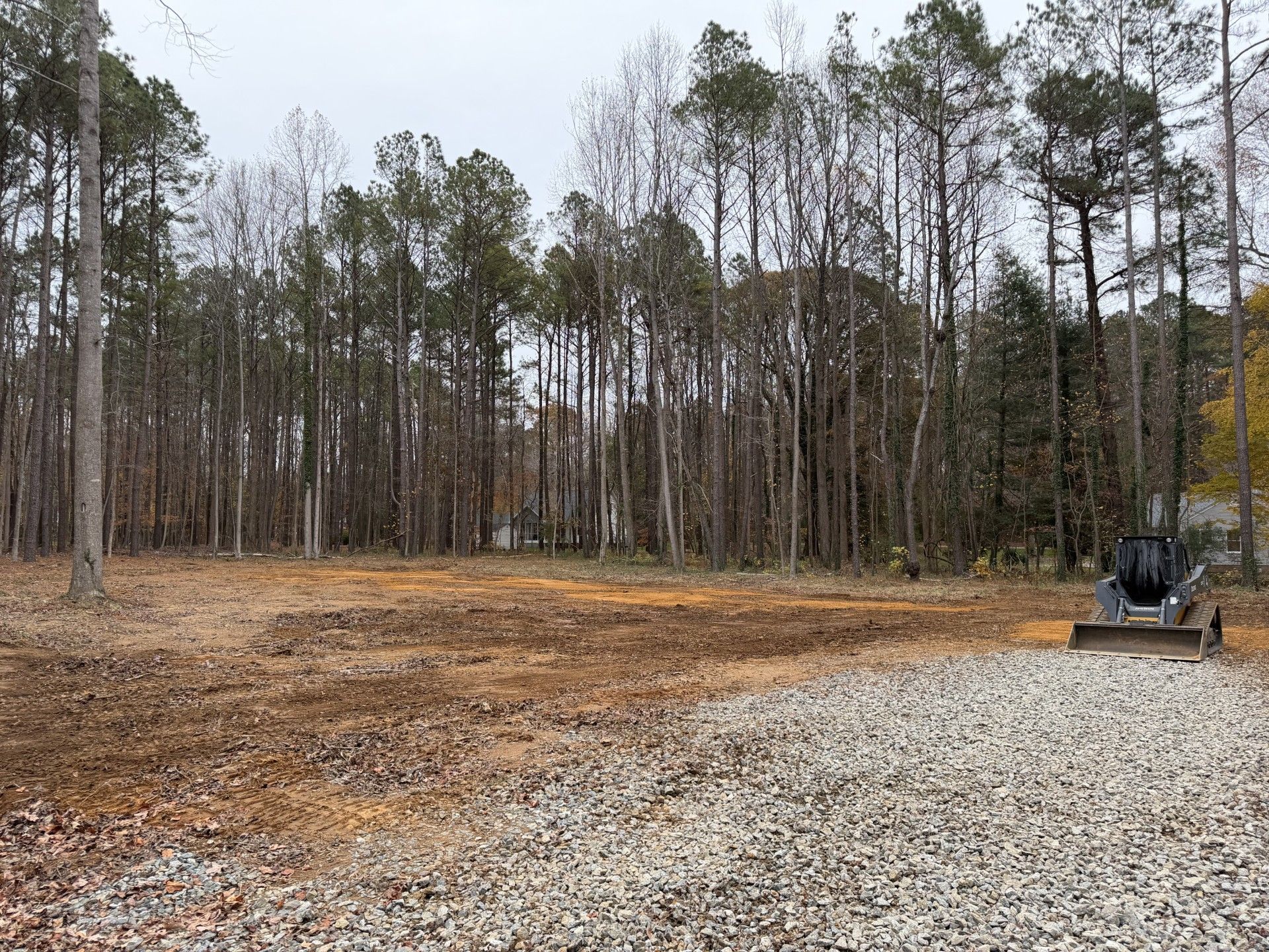 A cleared dirt area with gravel, a small tractor, and a forest in the background on an overcast day.