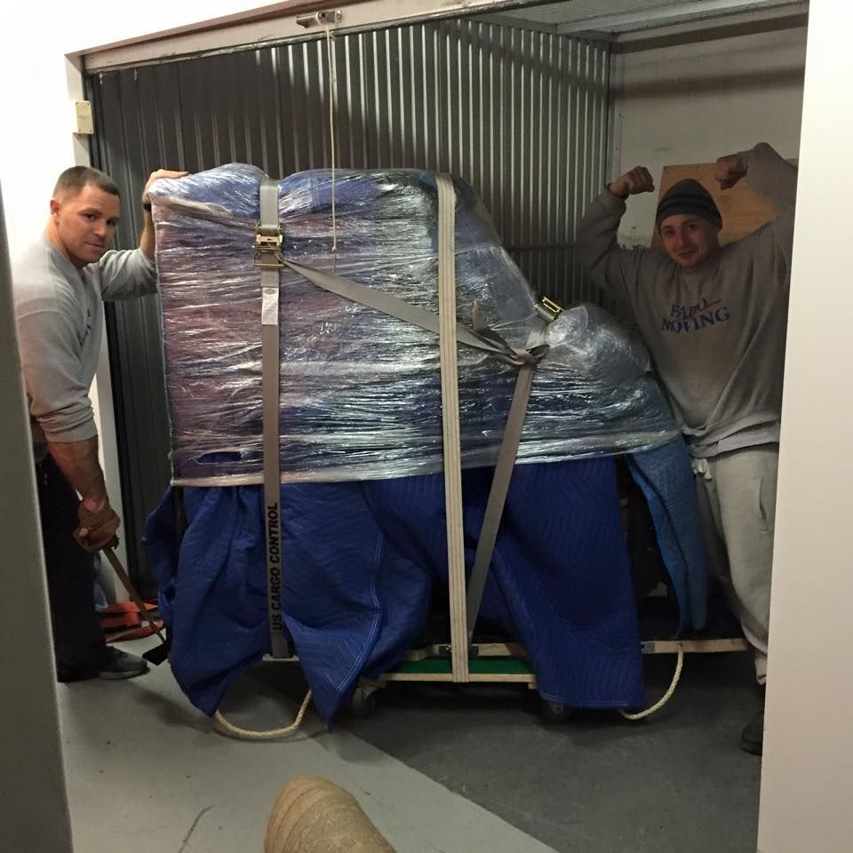 Two men moving a wrapped piano into a storage unit, one flexing his bicep.