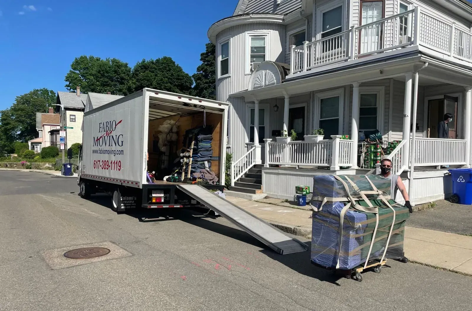 Moving truck unloading furniture from a house; blue, white, and brown colors; sunny day.