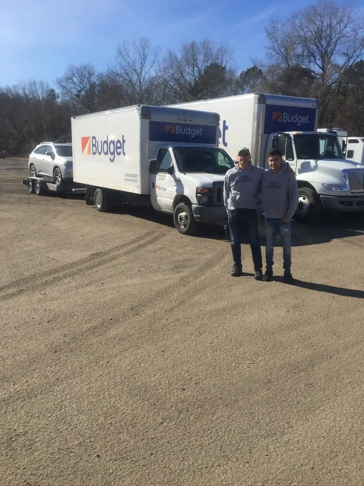 Two men stand by Budget moving trucks on a gravel lot on a sunny day. A car is on a trailer.