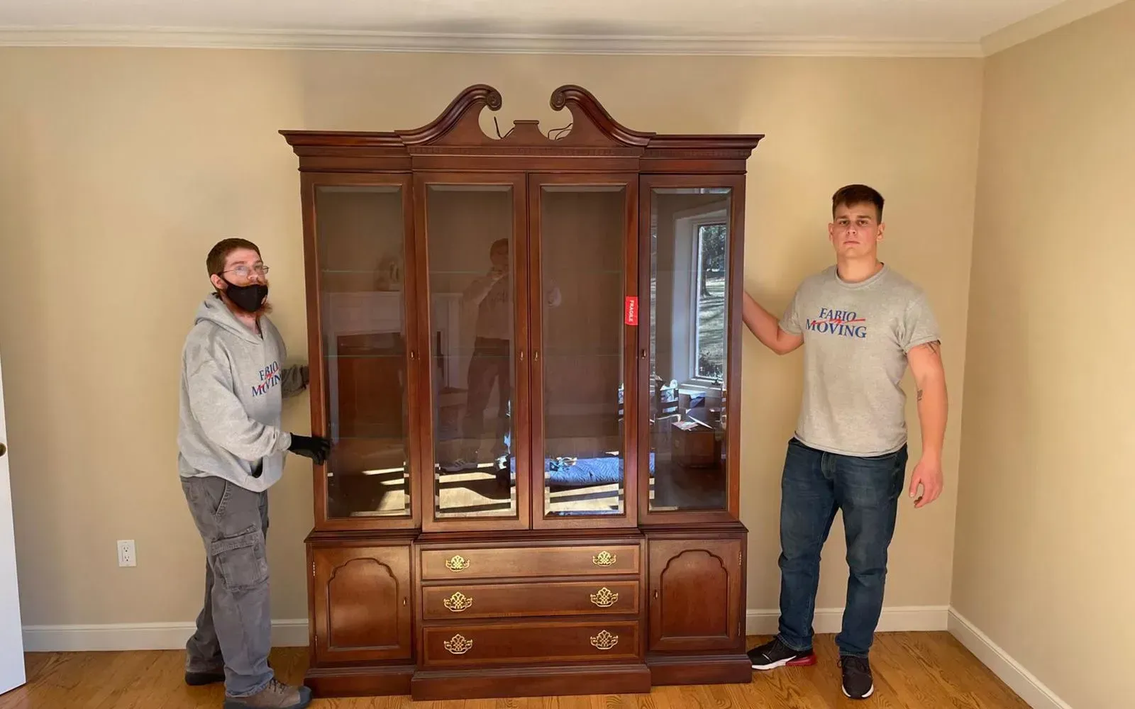 Two men moving a large, dark wood cabinet with glass doors in a room with tan walls.