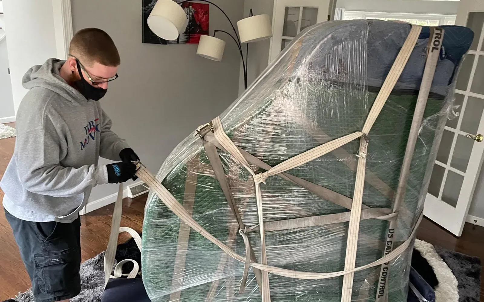 Man in mask straps a wrapped chair indoors, preparing for a move.