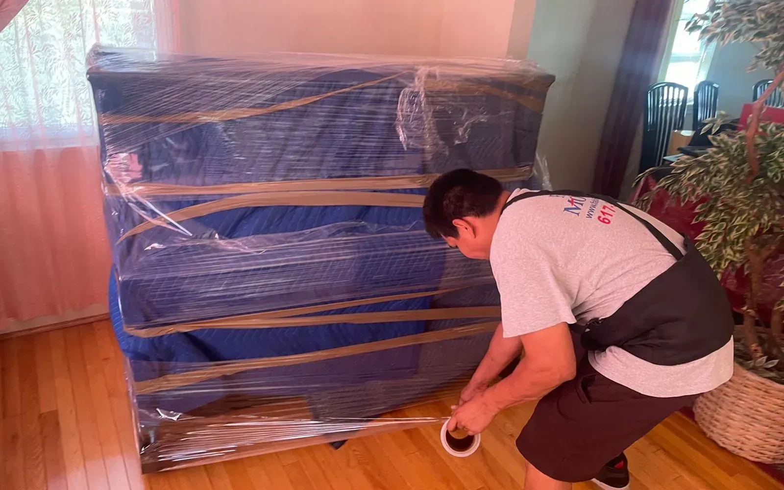 Man wrapping a blue, wrapped furniture with clear plastic and tape. Interior shot.