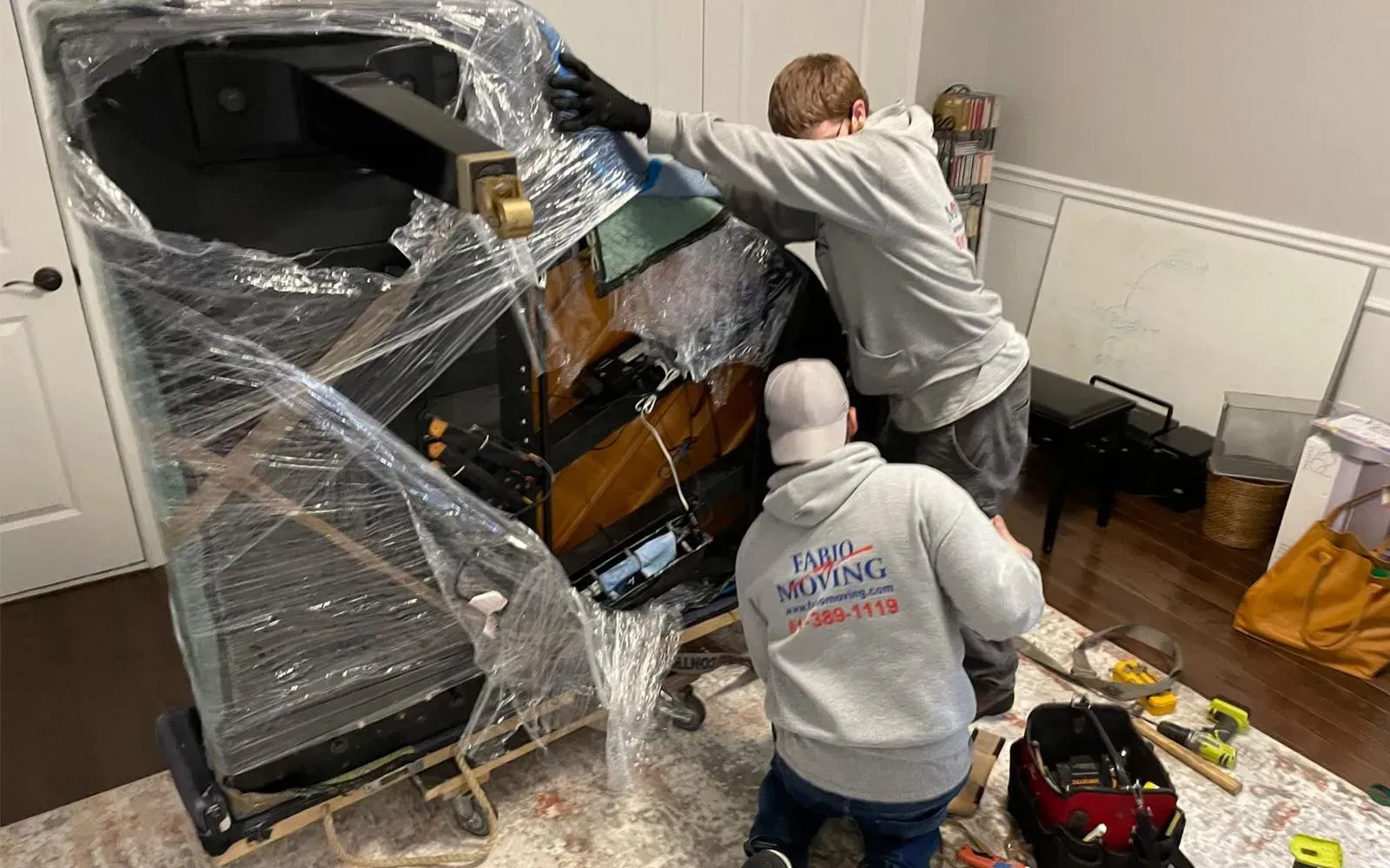 Two men moving a wrapped piano. Indoors, they wear gray hoodies, one kneels, and tools are visible.