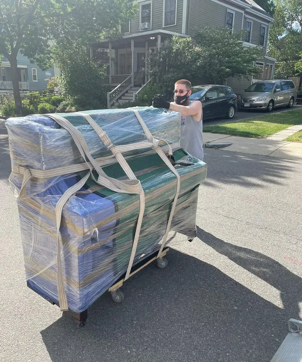 A man moving wrapped furniture on a dolly in a sunny street.
