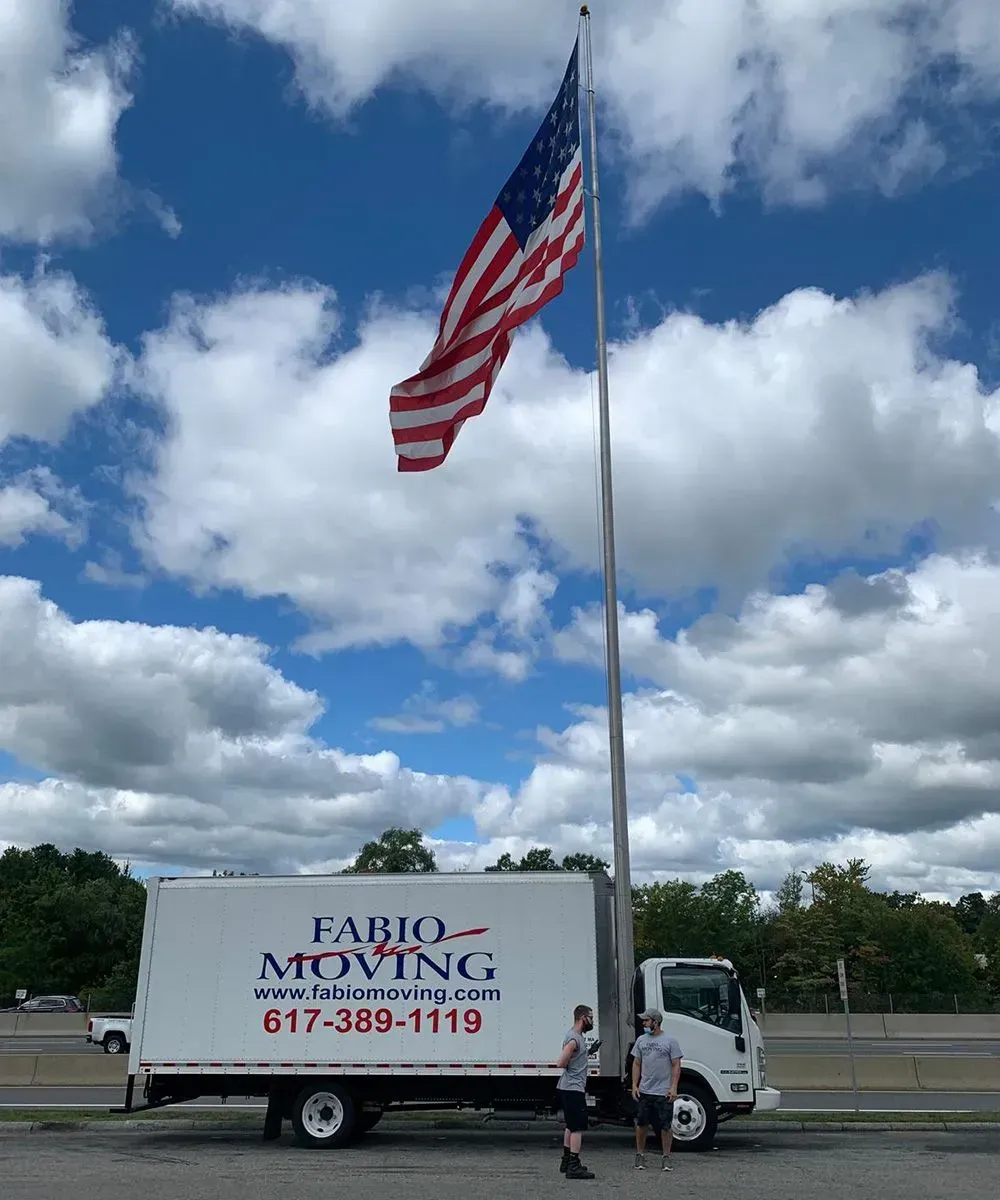 Two men stand beside a Fabio Moving truck beneath a tall flagpole with an American flag. Bright sky.