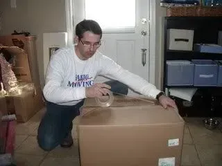 Man kneels to tape a cardboard box, indoors. He wears glasses and a long-sleeved shirt.