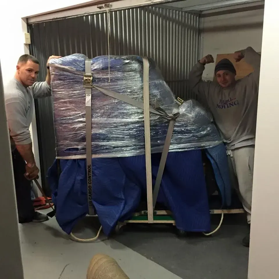 Two men loading a wrapped blue piano into a storage unit, one with a raised arm, showing effort.