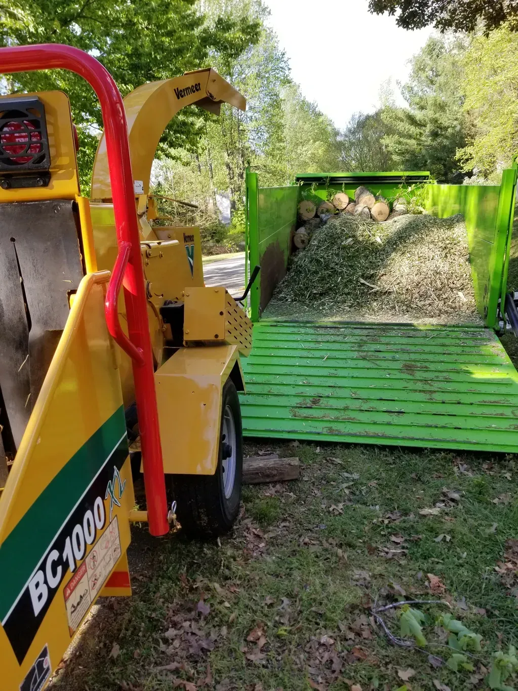 A yellow chipper is sitting next to a green trailer filled with leaves.