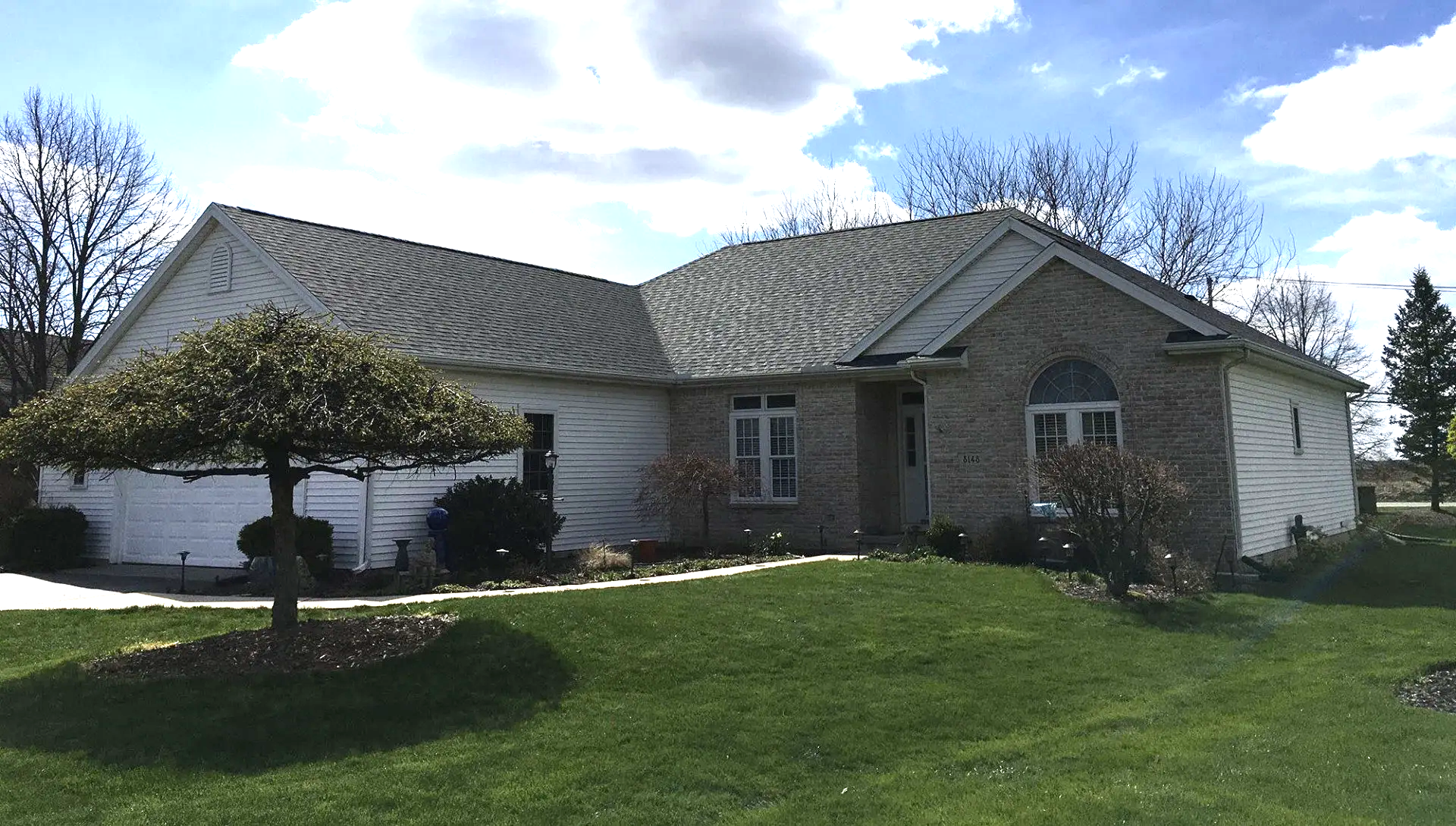Ranch-style house with brick and white siding, green lawn, and a small tree in front. Overcast sky.