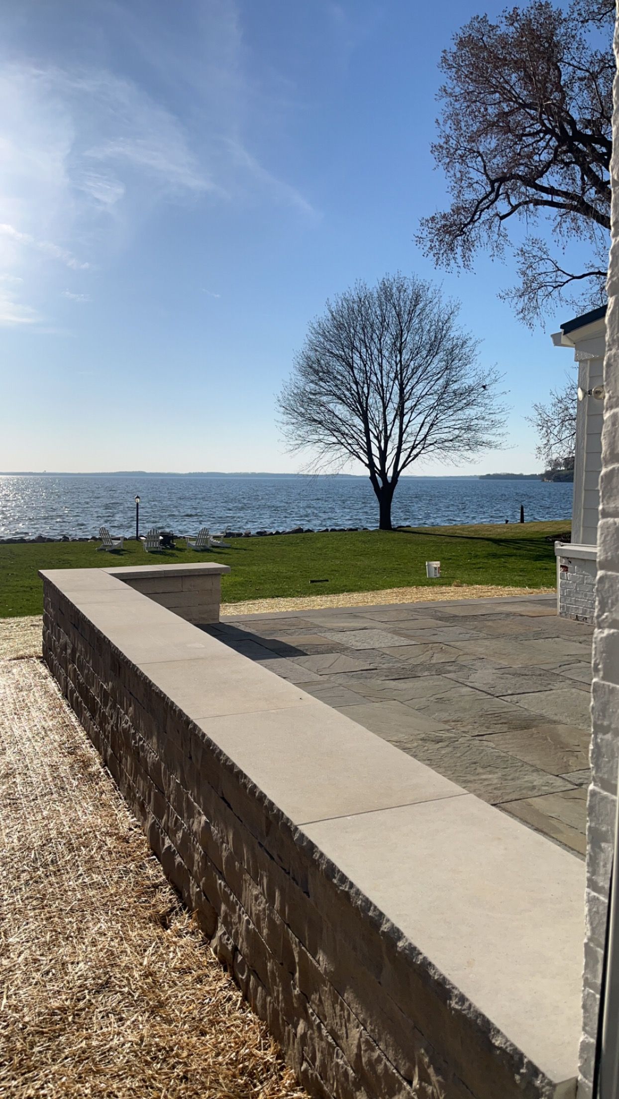 A concrete wall overlooks a grassy area, a tree, and a shimmering body of water under a sunny sky.