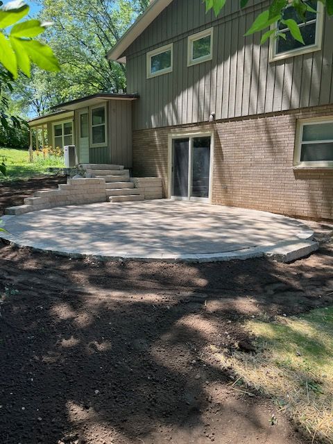 Circular stone patio next to a house with brick and siding. Dirt and grass surround the patio.