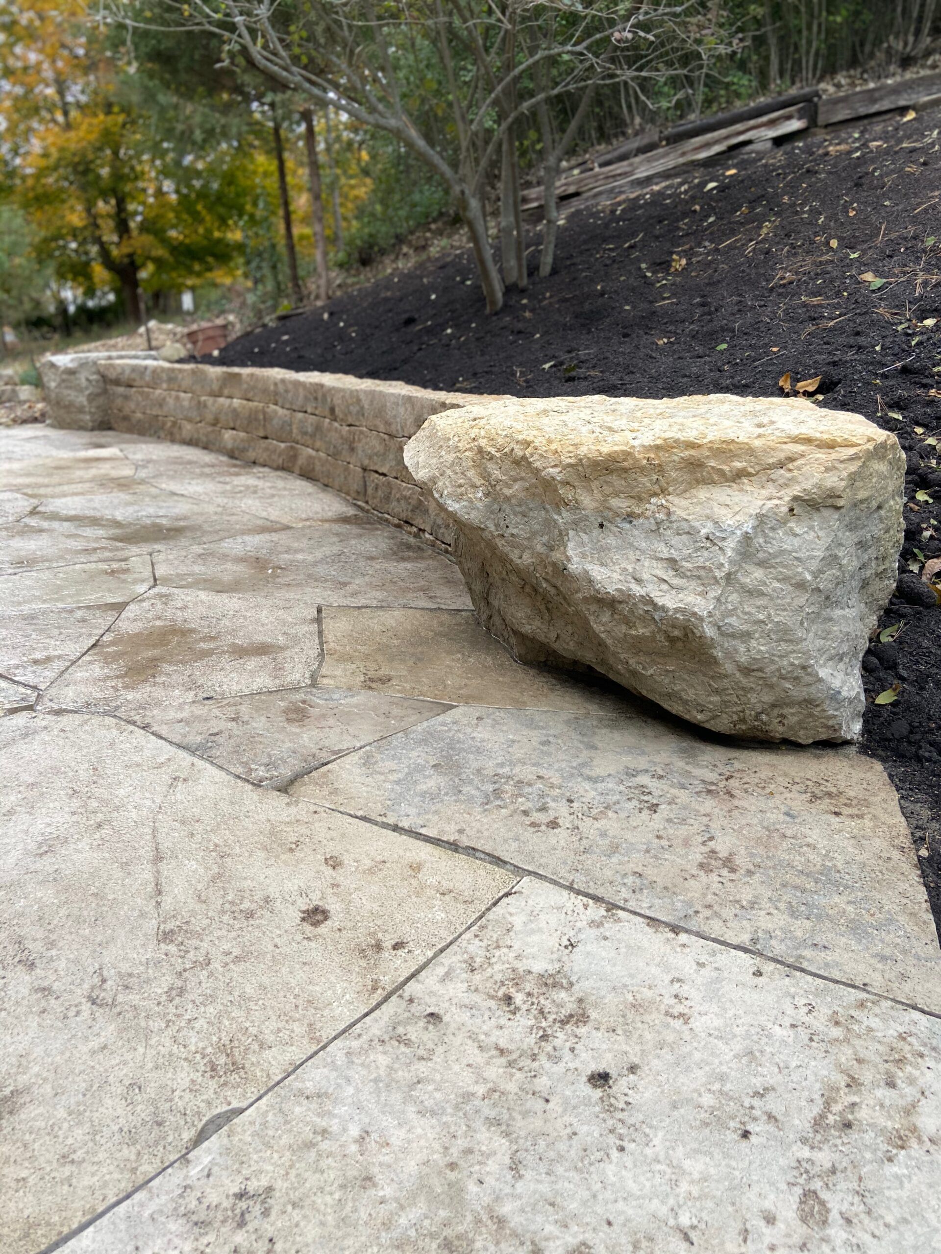 Stone wall with large rock on a paved area, hillside in the background.