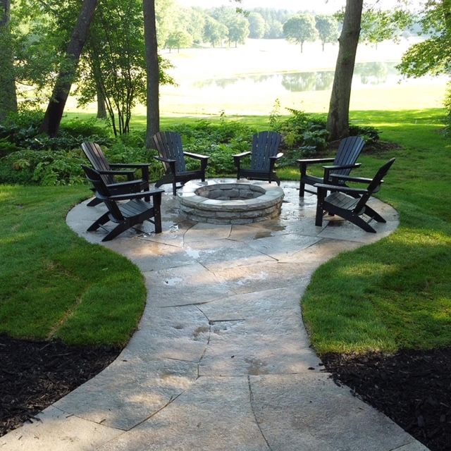 Stone fire pit surrounded by black Adirondack chairs on a stone patio, lawn in background.