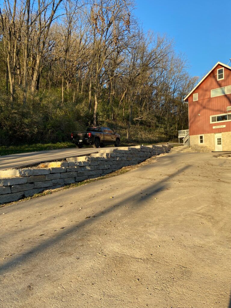 Gravel driveway with a truck parked near a red barn and stone retaining wall. Trees in the background.