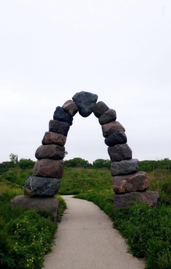 Stone archway over a paved path, leading into a green landscape under an overcast sky.