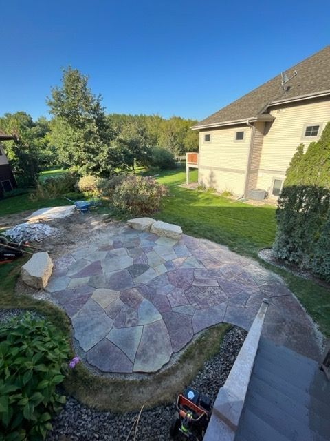 Stone patio with pathway next to a beige house, lawn, and trees under a blue sky.