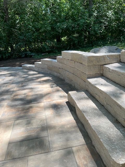 Stone steps and curved retaining wall built on a paved patio, with a treeline backdrop.