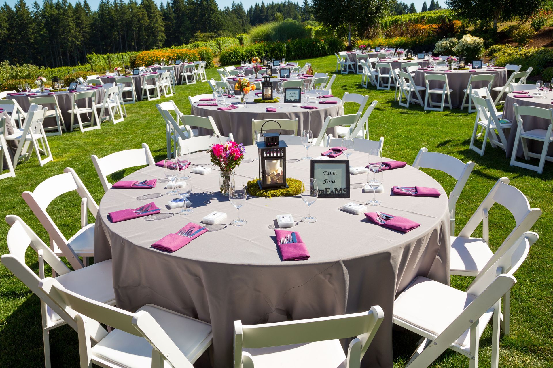 Outdoor reception with round tables set with gray linens and pink napkins, surrounded by white chairs on a grassy hill.