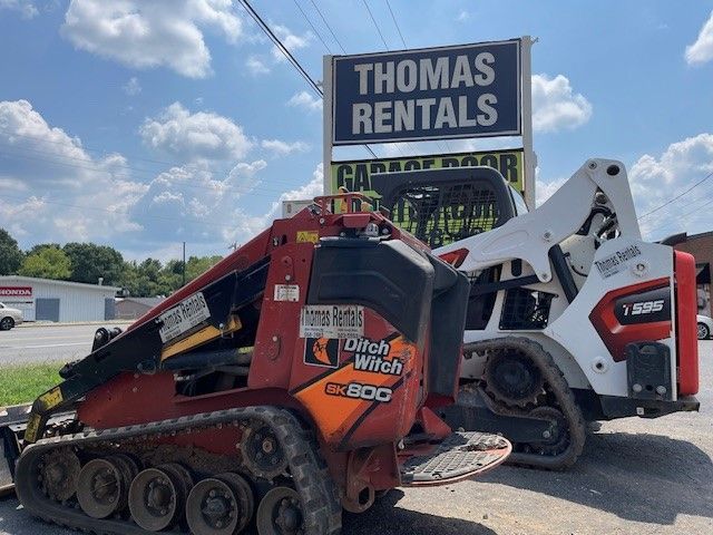 Two track loaders parked in front of Thomas Rentals sign. Red and white machines on a sunny day.