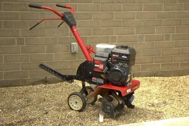 Red and black Troy-Bilt garden tiller on gravel against a brick wall.