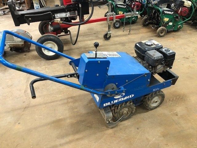 Blue self-propelled Bluebird lawn dethatcher with blue handles and engine. Located indoors on a concrete floor.
